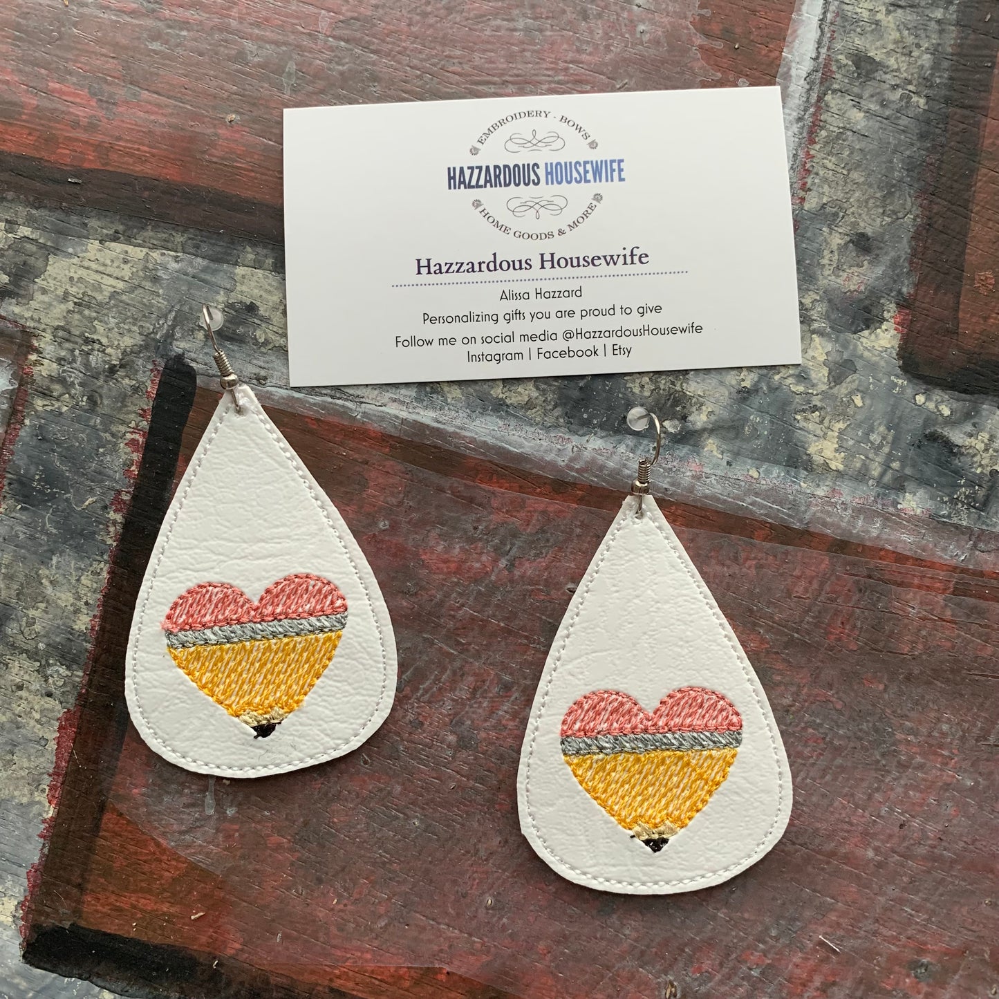 Teardrop-shaped earrings with embroidered pencil in the shape of a heart design on a brick surface with a business card.