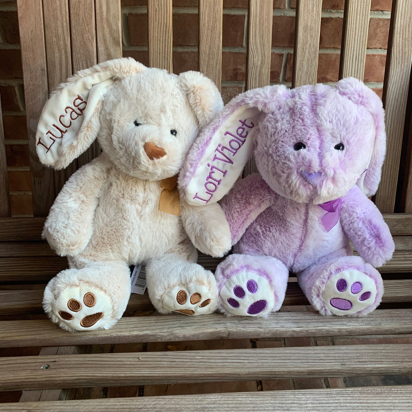 Two plush toys, one beige and one purple with embroidered names on ears, sitting on a wooden surface