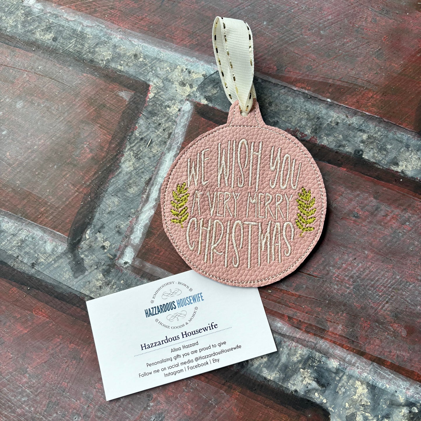 Pink Christmas ornament with 'Merry Christmas' text on a wooden surface, accompanied by a HazzardousHousewife business card.