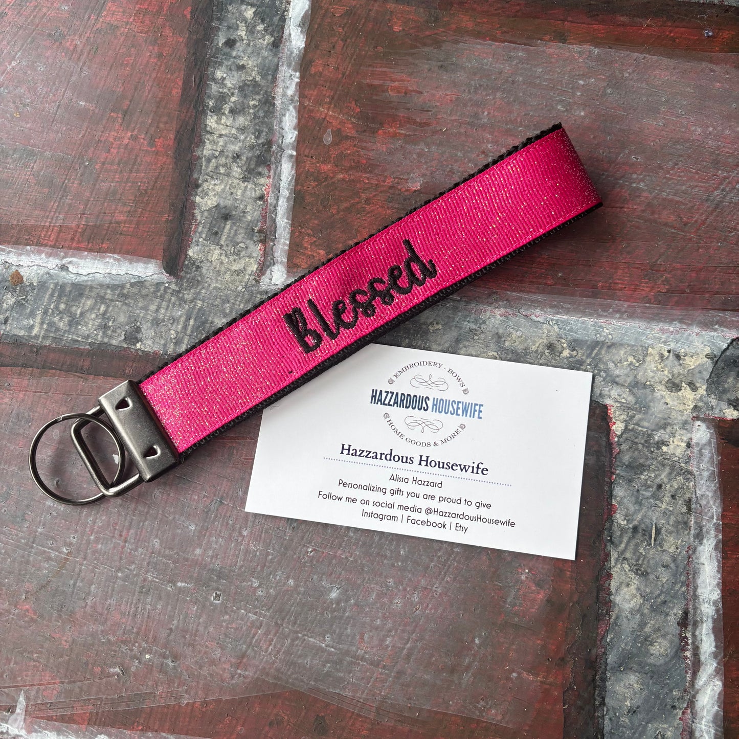 Pink keyfob with black webbing showing at the edge with 'Blessed' embroidered in black text on a wooden surface
