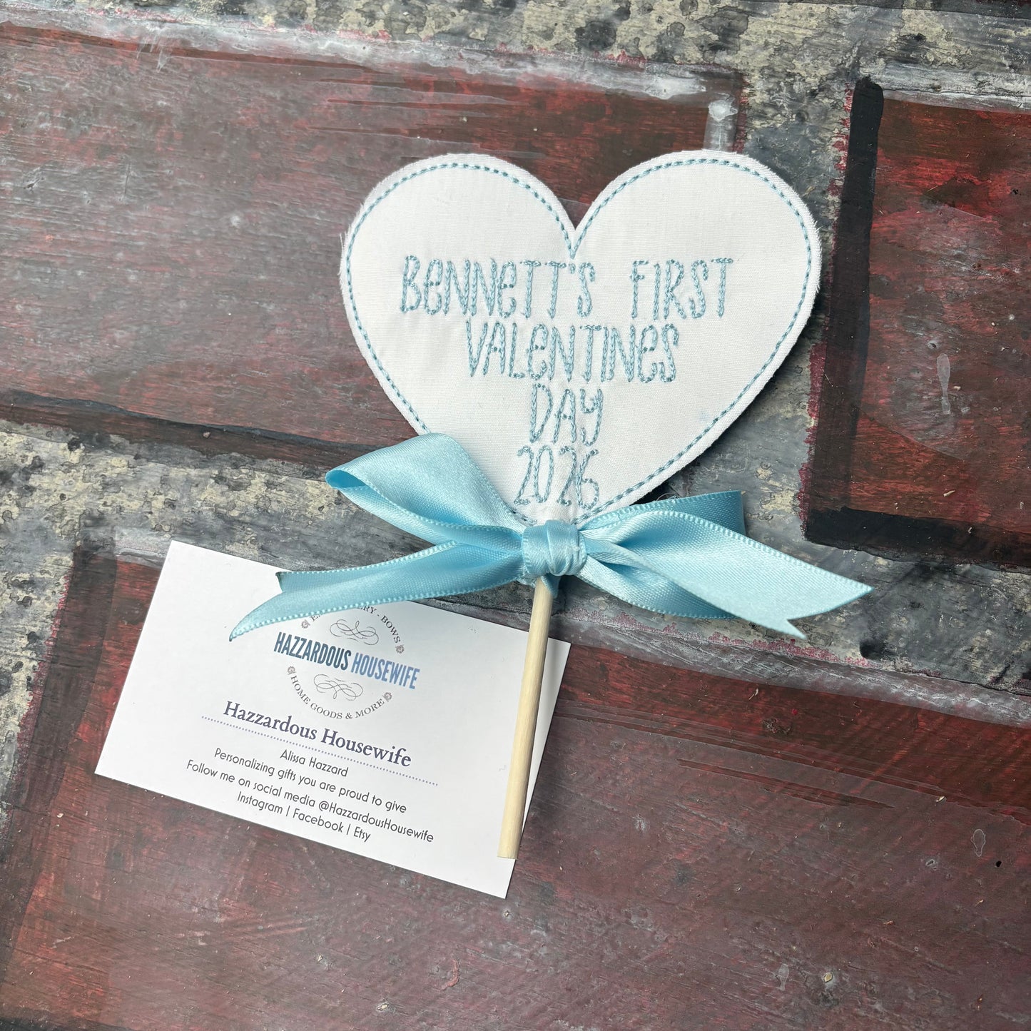 Heart-shaped sign with 'Bennett's First Valentine's Day 2026' text and a blue ribbon on a rustic wooden background.