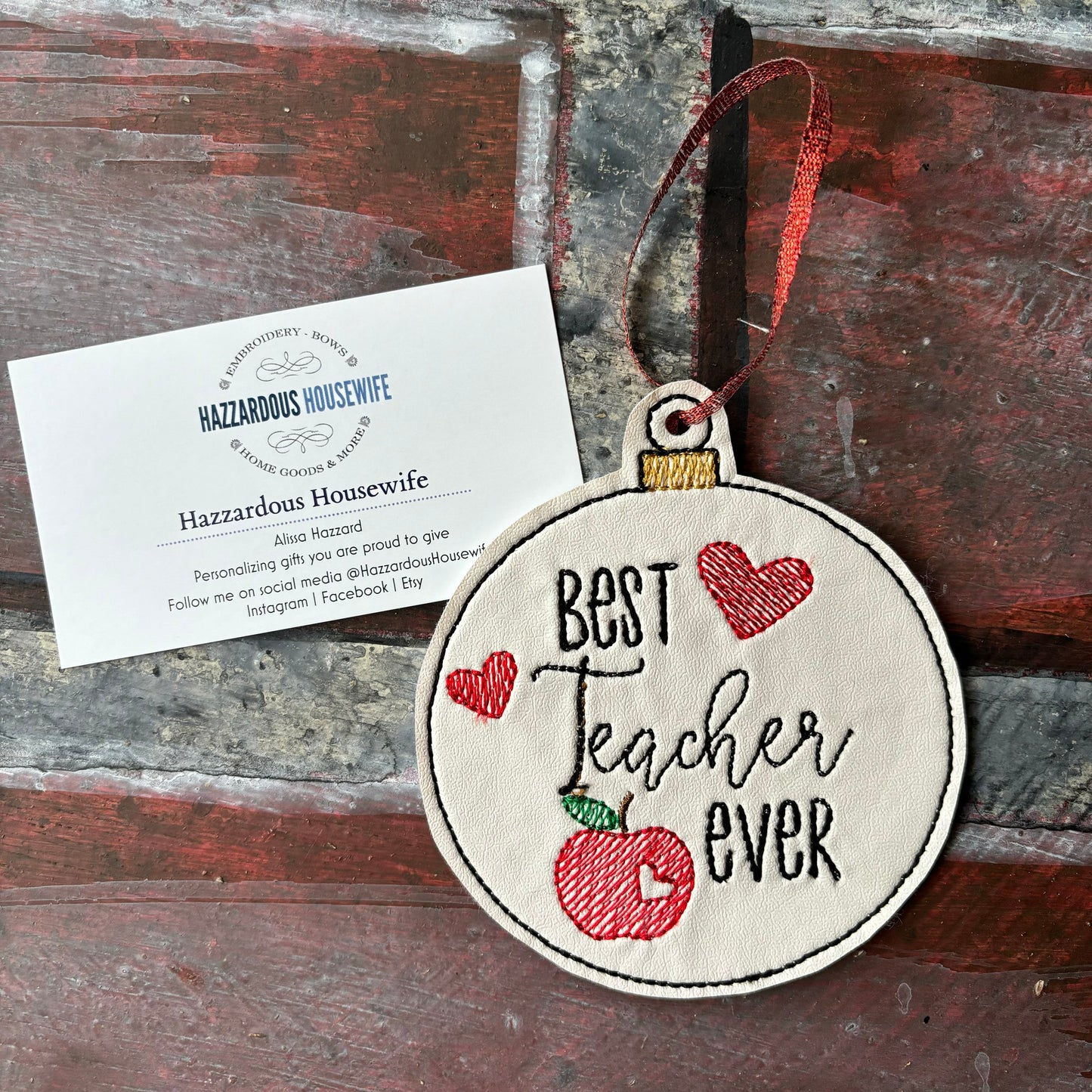 Decorative ornament with 'Best Teacher Ever' text and an apple, placed on a wooden surface with a business card.