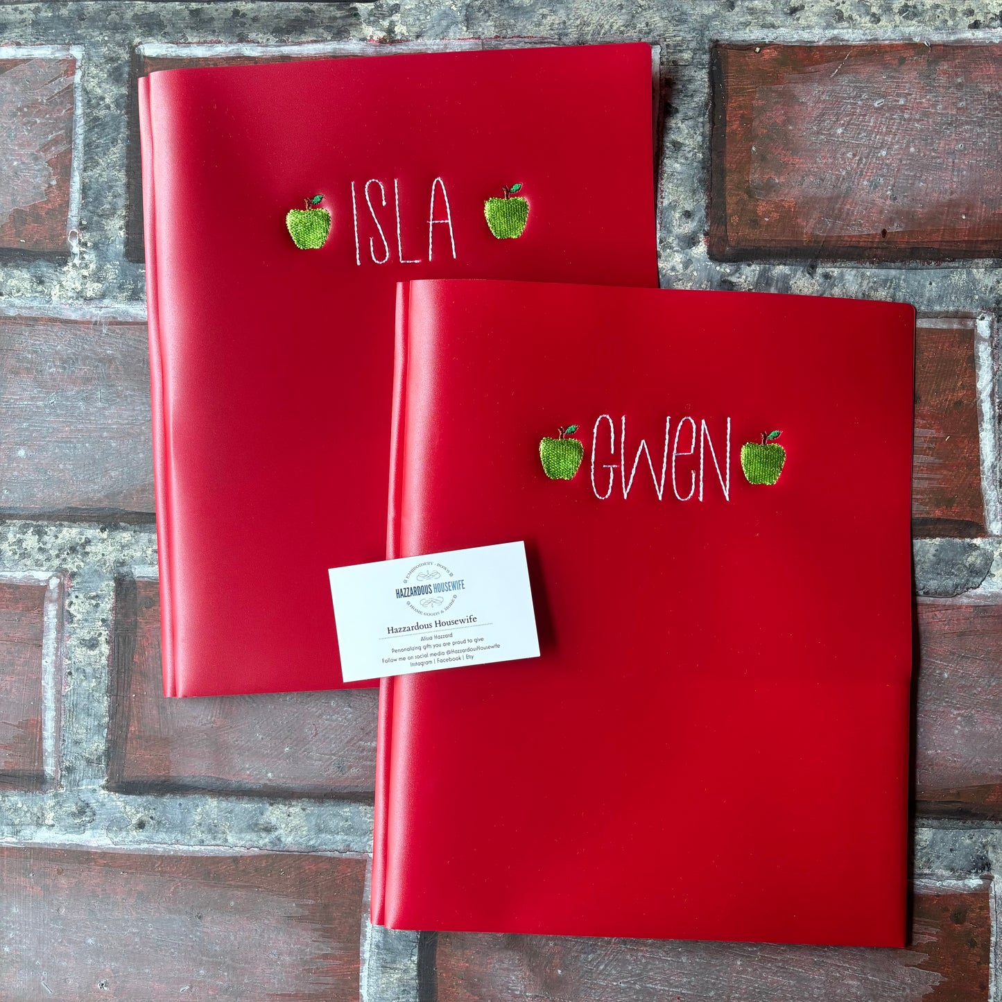 Two red plastic folders with embroidered names and green apples on a brick surface
