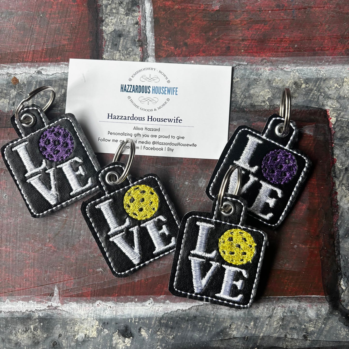 Four 'LOVE' pickleball keychains with colorful centers on a textured surface, featuring a card with 'Hazzardous Housewife' branding.