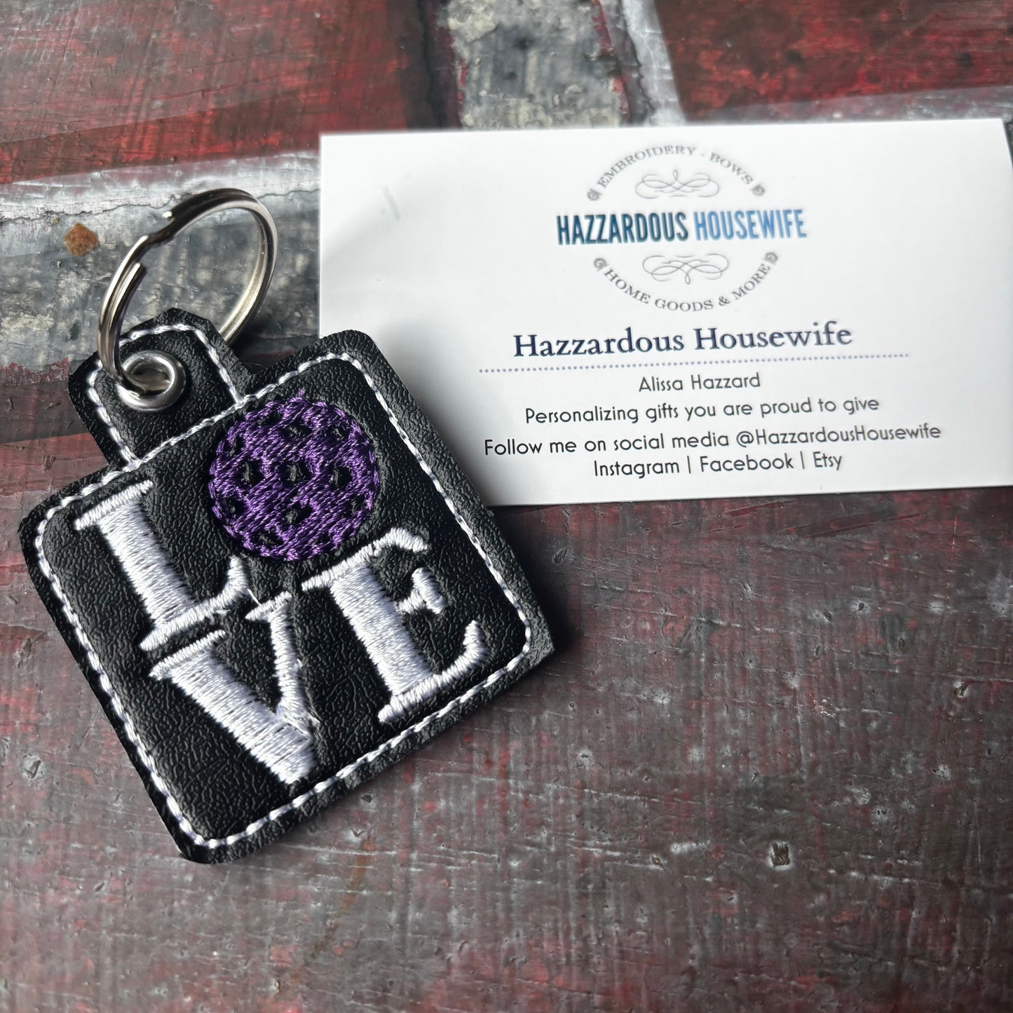 Pickleball Keychain with 'LOVE' embroidery and purple pickleball ball on a business card of 'Hazzardous Housewife' brand.