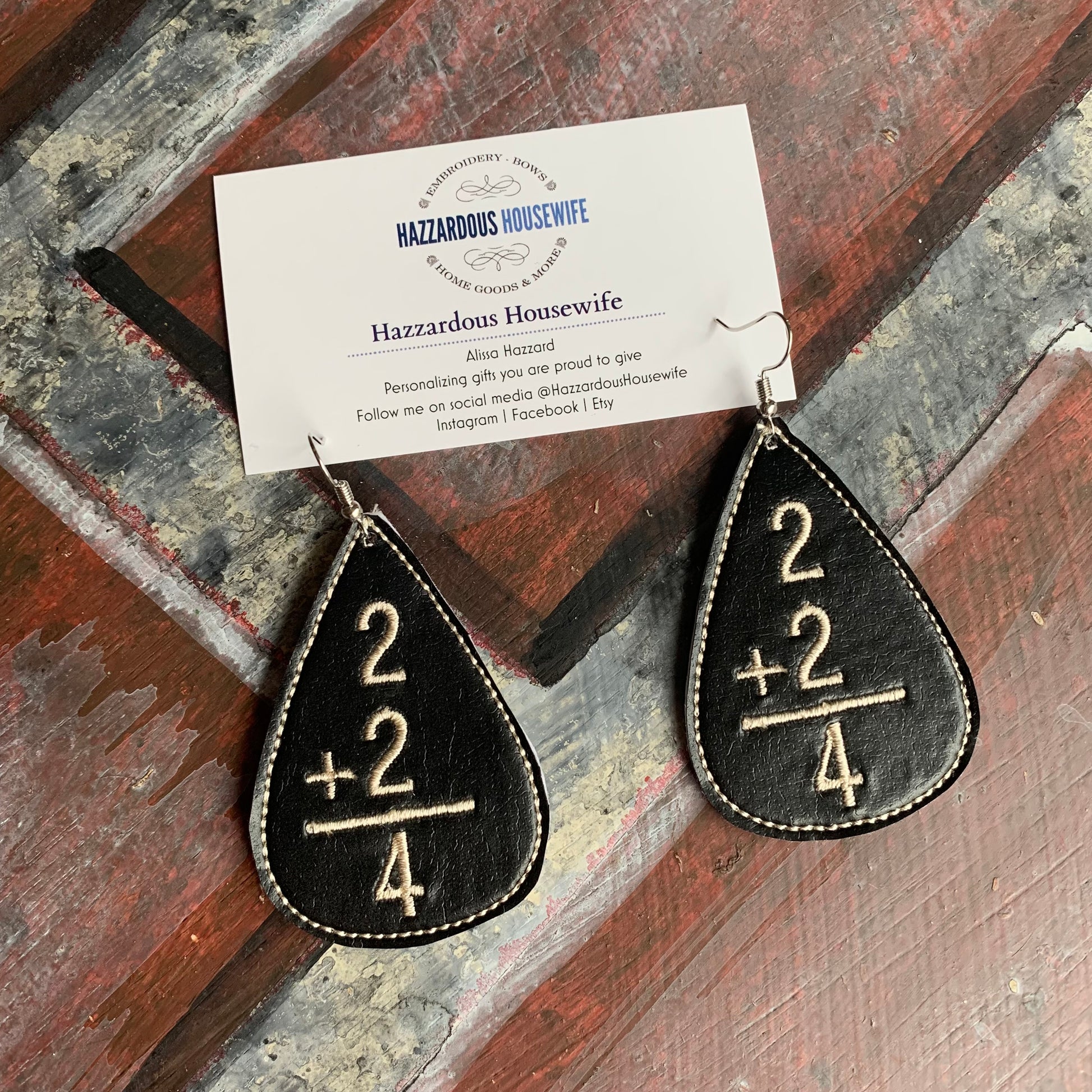 Black teardrop earrings with mathematical design on a wooden surface, featuring 'Hazzardous Housewife' branding.