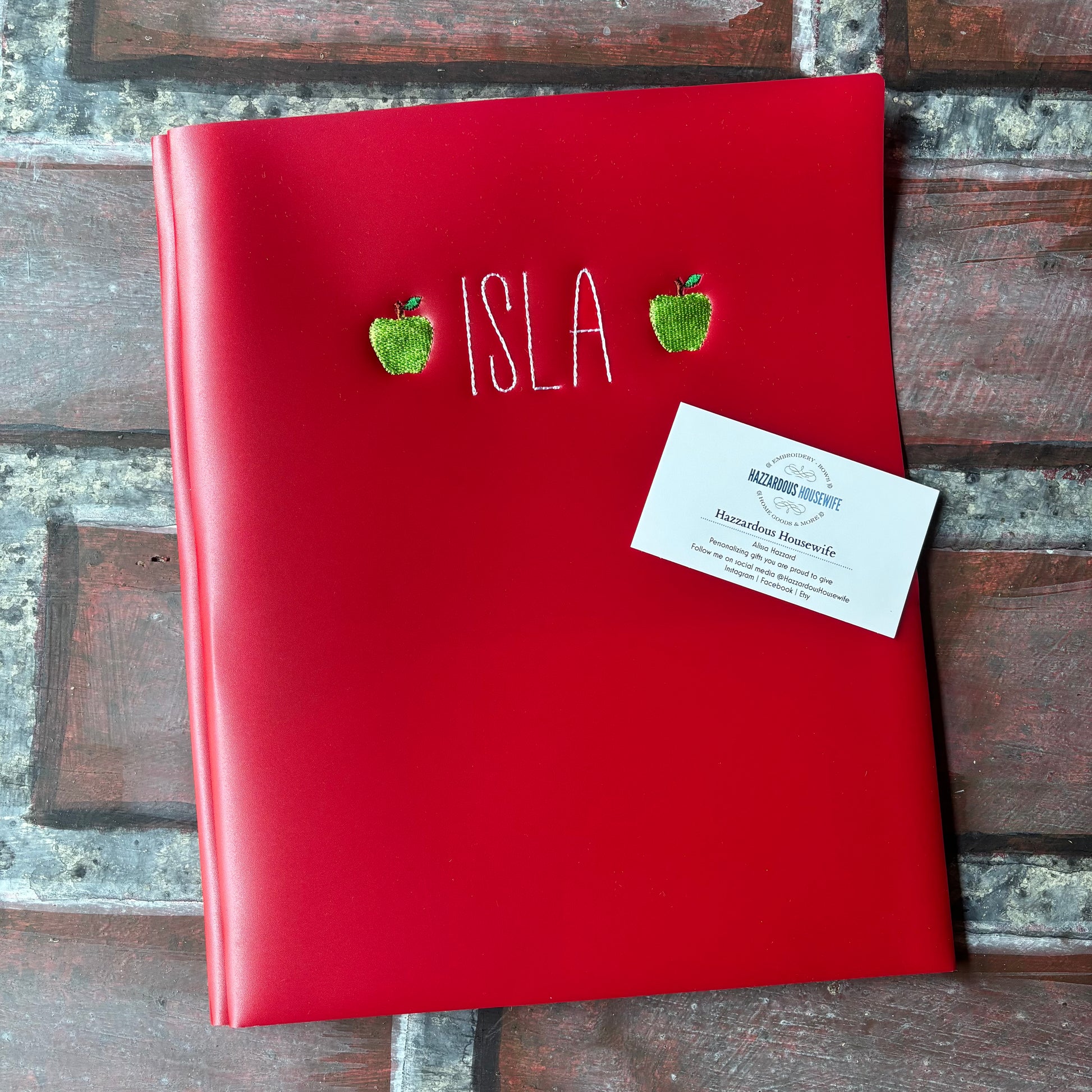 Red plastic folder with 'ISLA' embroidered on it and two green apples, placed on a brick surface.