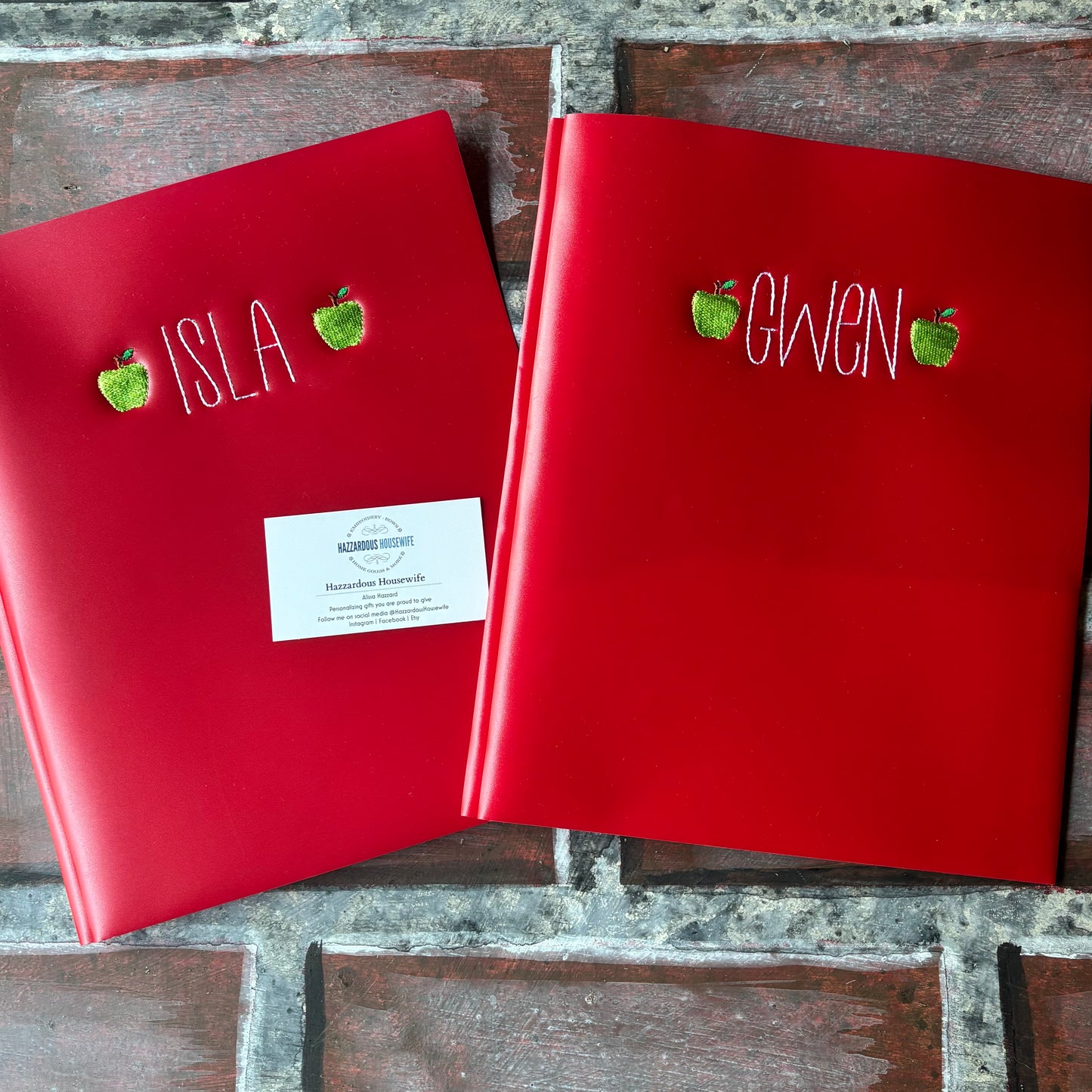 Two red plastic folders with embroidered names and green apples on a brick surface