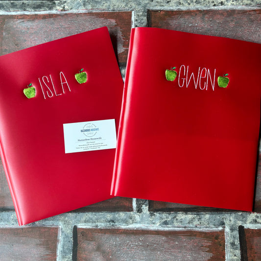 Two red plastic folders with embroidered names and green apples on a brick surface