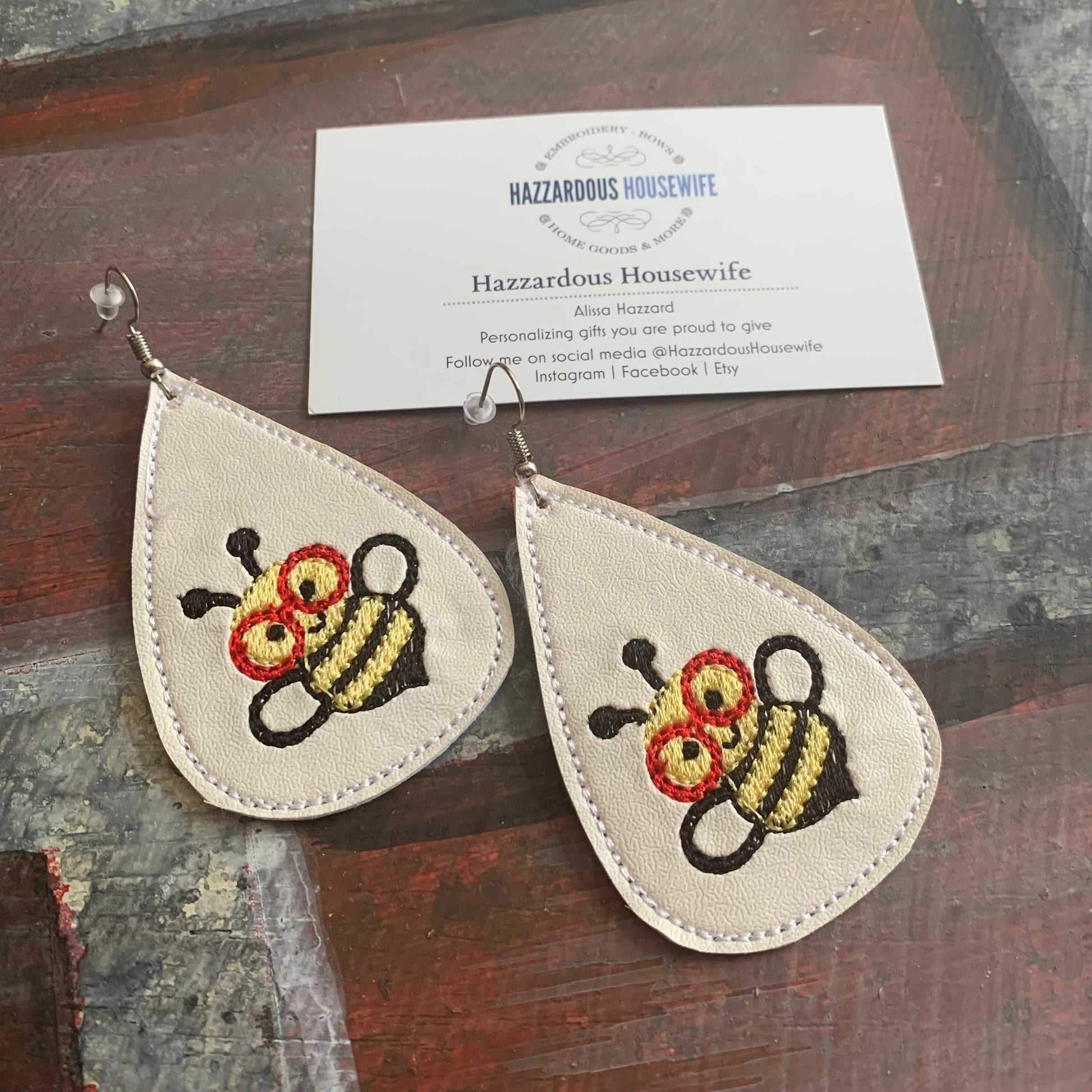 Teardrop-shaped earrings with embroidered bee wearing red glasses design on a wooden surface with a business card.
