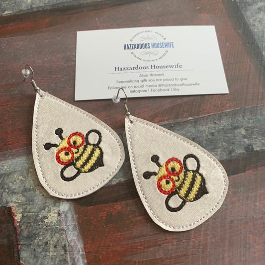 Teardrop-shaped earrings with embroidered bee wearing red glasses design on a wooden surface with a business card.