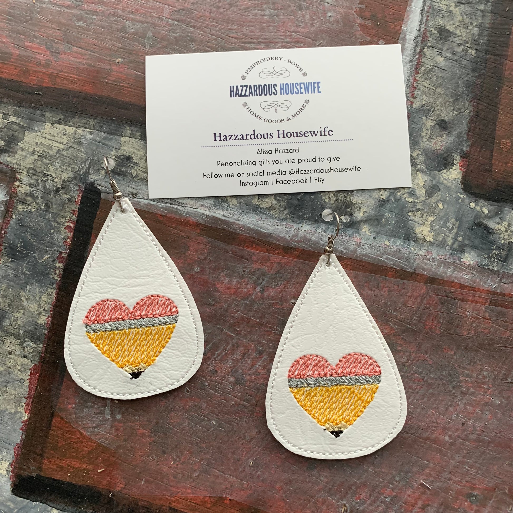 Teardrop-shaped earrings with embroidered pencil in the shape of a heart design on a brick surface with a business card.
