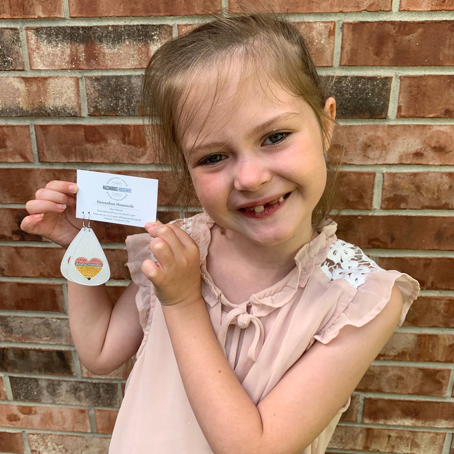 Young girl holding a business card and heart-shaped pencil embroidered on teardrop earring against a brick wall