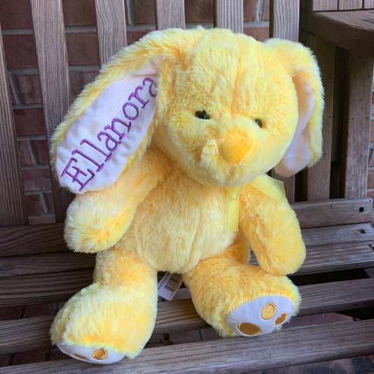 Yellow stuffed bunny with an embroidered name on the ear in purple font on a wooden bench