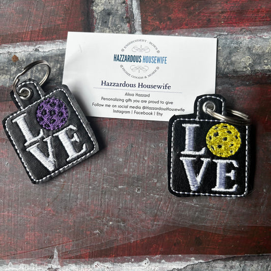 Two 'LOVE' keychains with colorful centers on a wooden surface with a business card. The O is a yellow or purple pickleball.