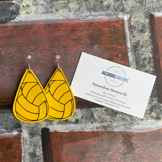 Yellow earrings with black patterns on a rustic wooden surface next to a business card.