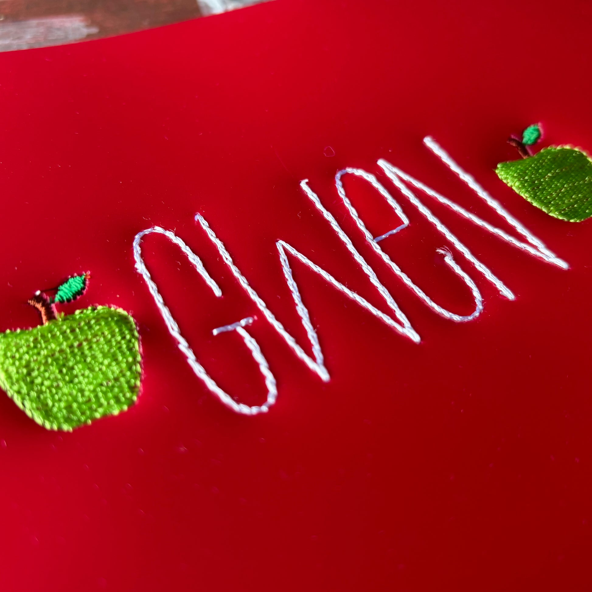 Embroidered name 'Gwen' on a red plastic folder with stitched green apples.