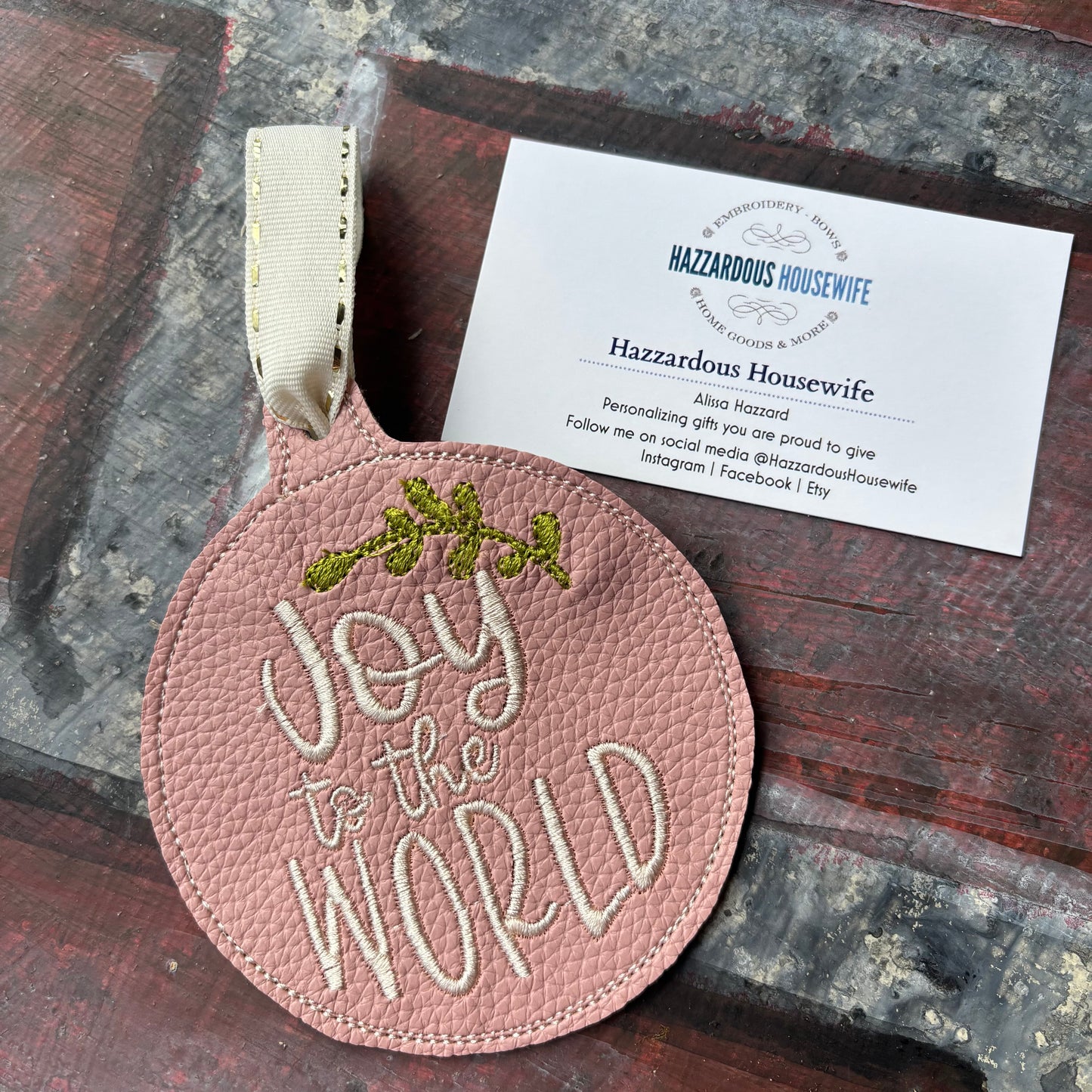 Pink leather ornament with 'Joy to the World' embroidery on a rustic surface with a business card.