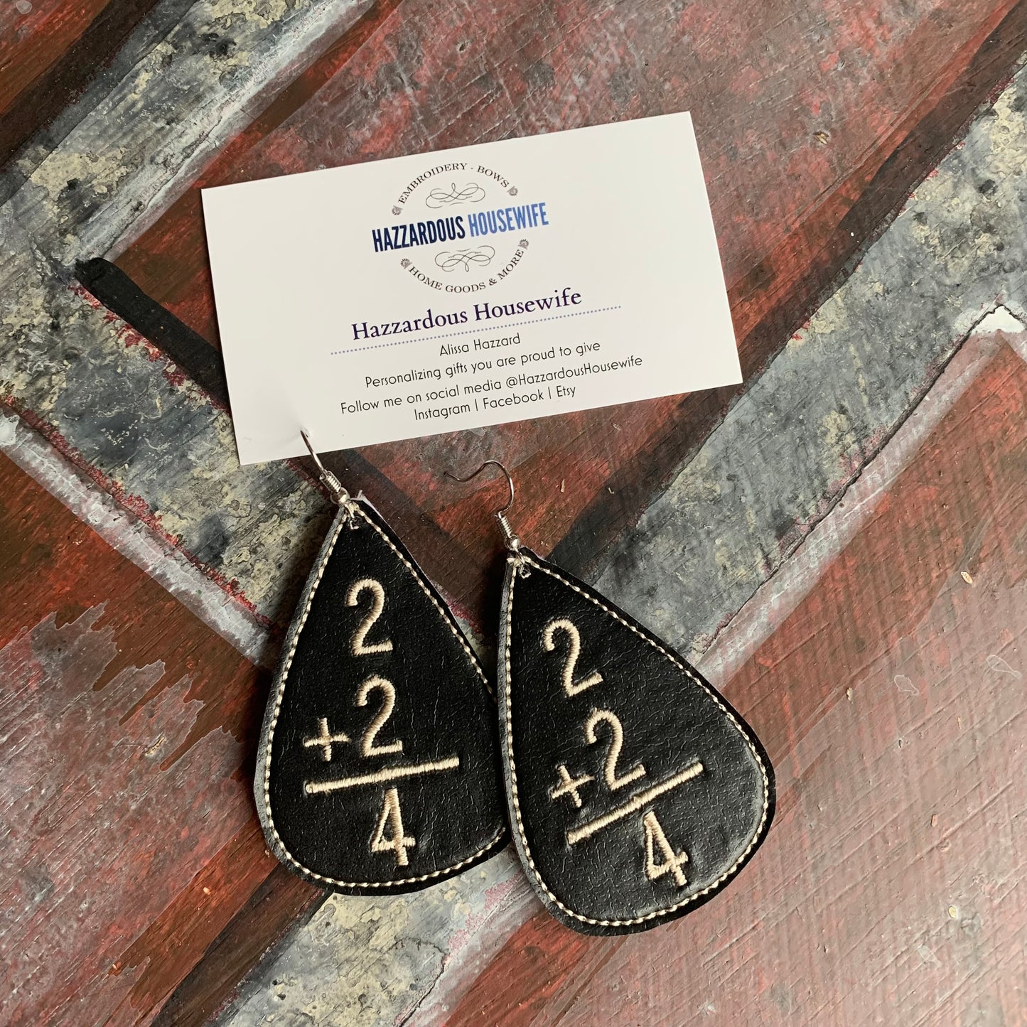 Teardrop-shaped earrings with mathematical design on a wooden surface with a business card.