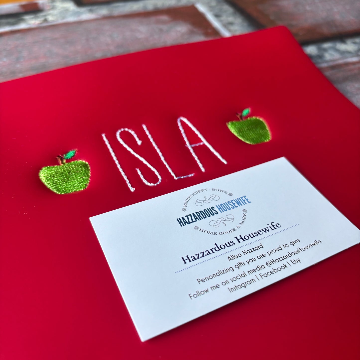 Red folder with 'ISLA' and green apples, featuring a card from Hazzardous Housewife on a brick surface.