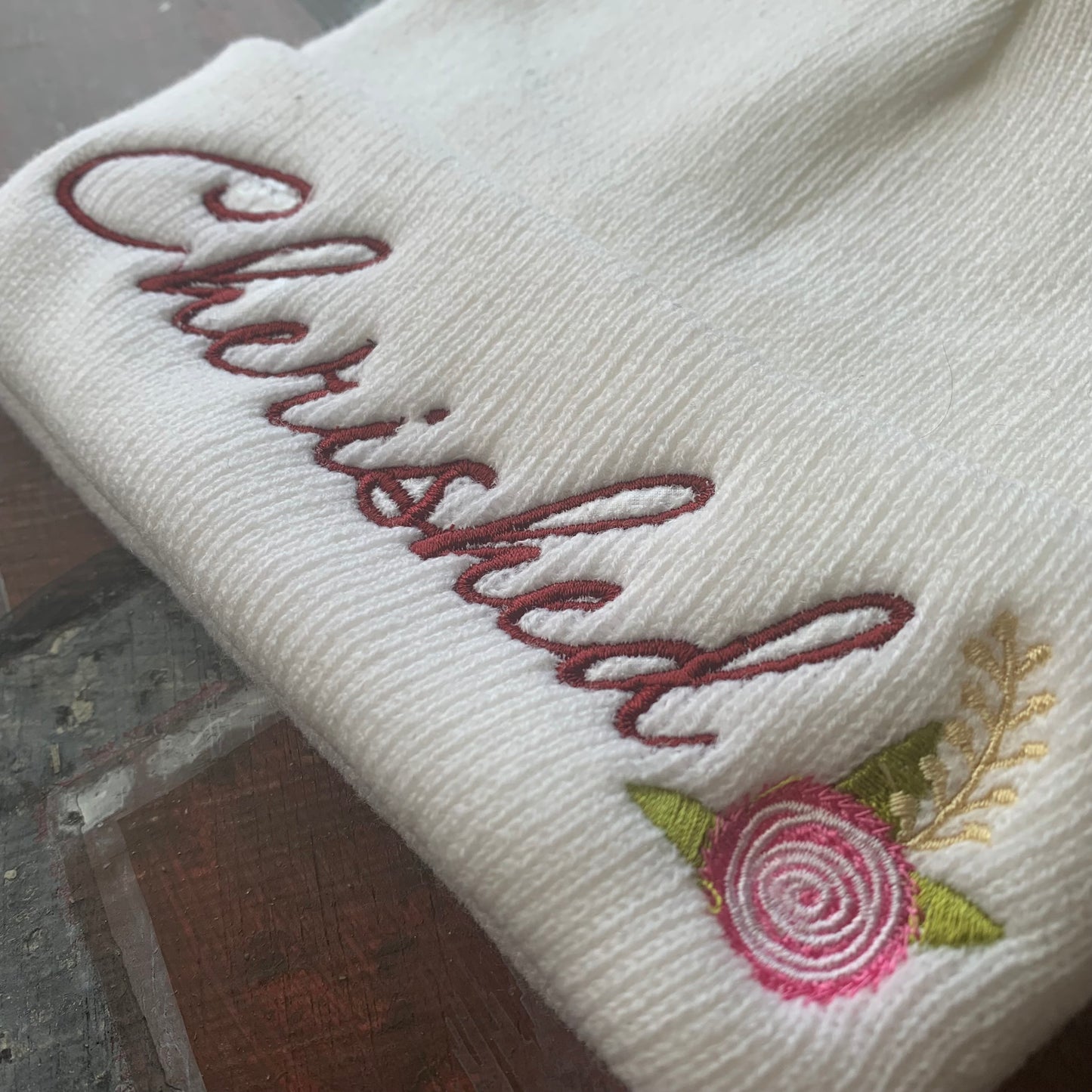 White beanie with 'Cherished' embroidery and floral design on a textured surface.