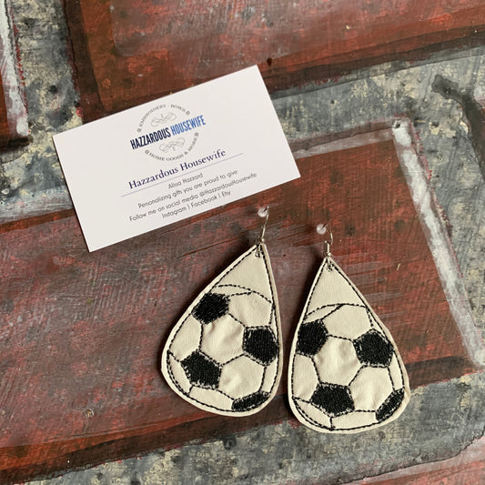 Two teardrop-shaped soccer ball patterned earrings on a rustic wooden surface with a HazzardousHousewife business card.