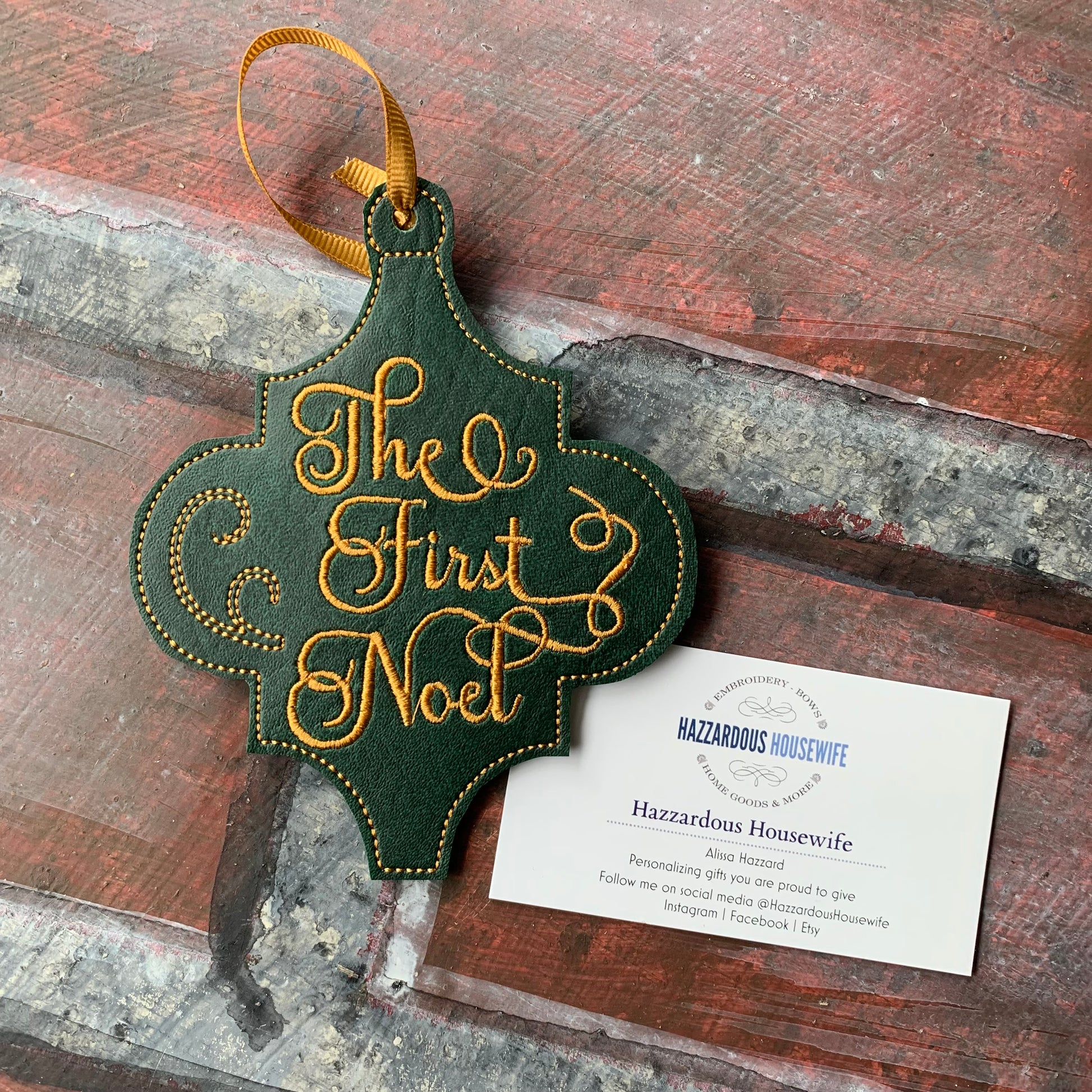 Decorative green ornament with 'The First Noel' text on a rustic wooden surface, featuring a card from Hazzardous Housewife.