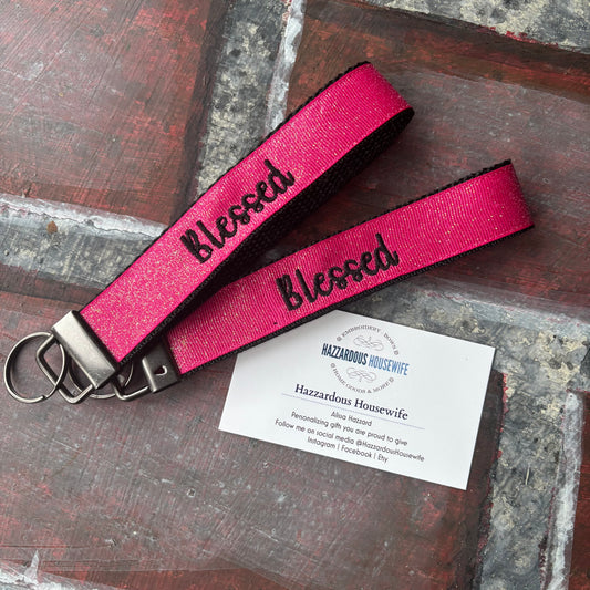 Pink keyfob with 'Blessed' text on a rustic wooden surface with a HazzardousHouswife business card.