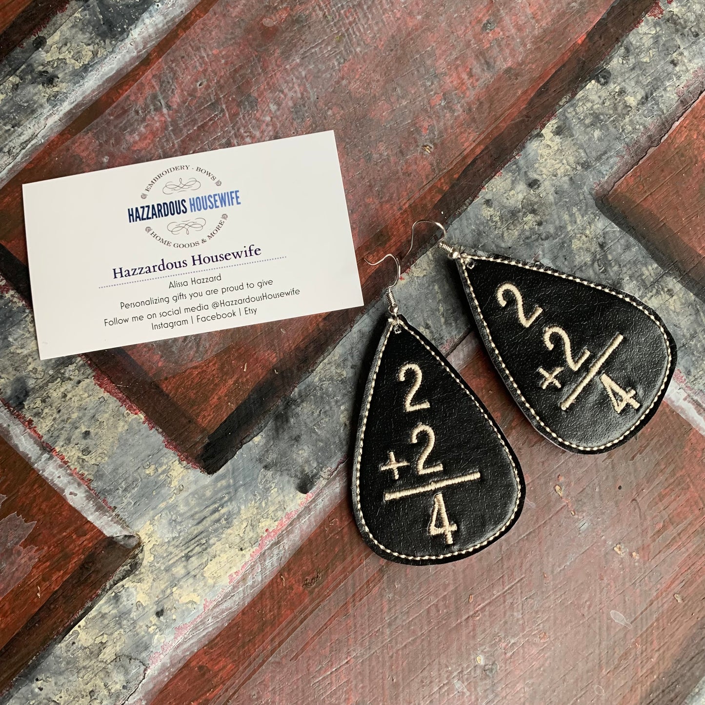 Black teardrop-shaped earrings with gold numbers on a wooden surface, accompanied by a business card for 'Hazzardous Housewife'.