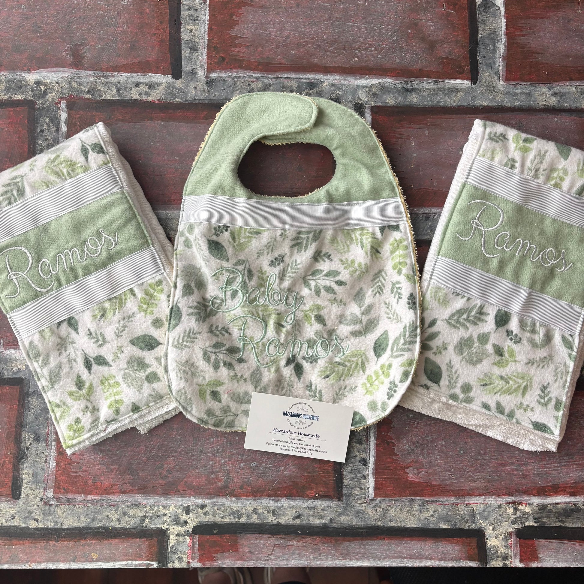 Set of nature green baby bib and burp cloths with personalized name branding on a brick surface.