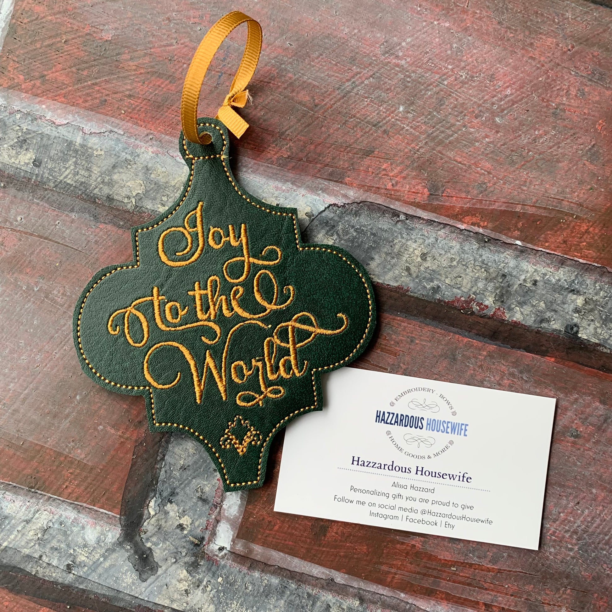 Decorative ornament with 'Joy to the World' text on a rustic wooden surface, featuring a HazzardousHousewife product card.