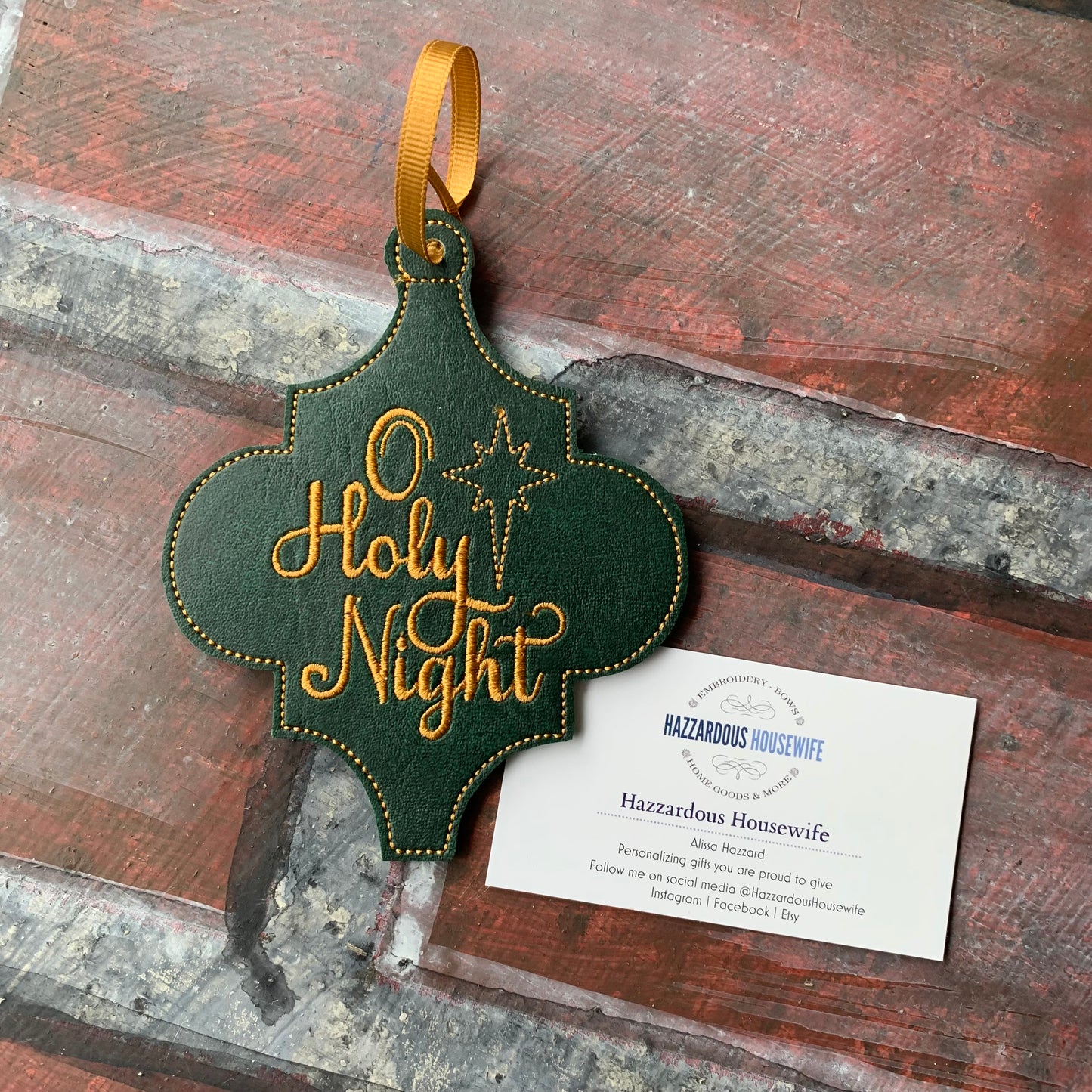Decorative green and gold 'O Holy Night' ornament on a rustic wooden surface with a HazzardousHousewife business card underneath.