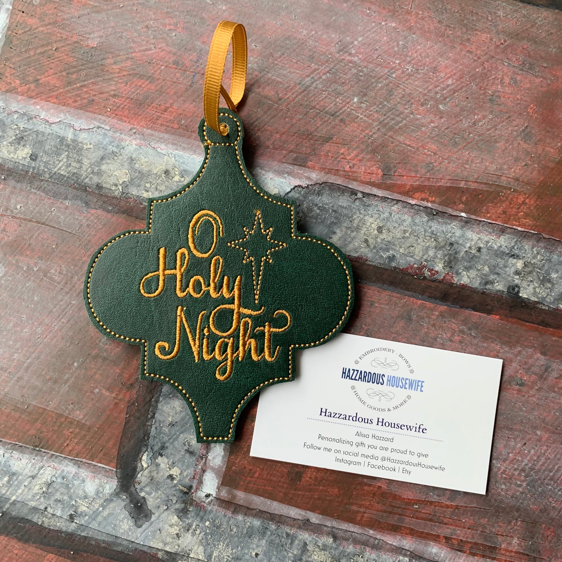 Decorative green and gold 'O Holy Night' ornament on a rustic wooden surface with a HazzardousHousewife business card underneath.
