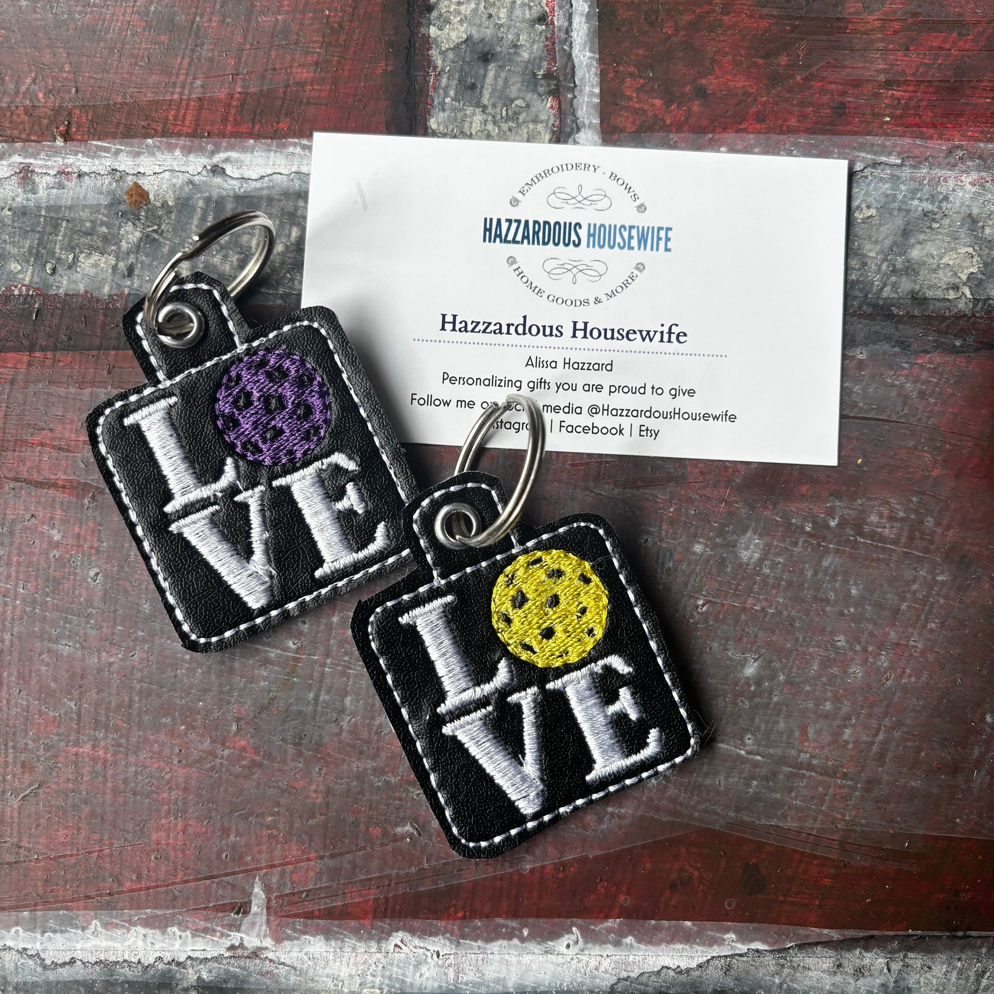 Two 'LOVE' pickleball keychains with colorful designs on a wooden surface with a business card.