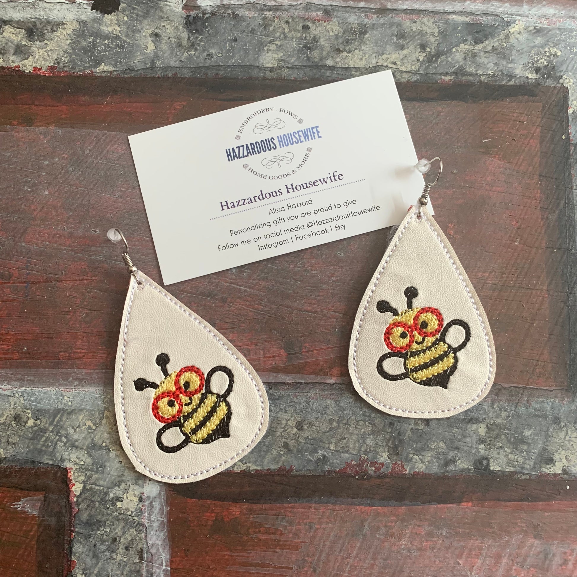 Teardrop-shaped earrings with embroidered bee designs on a rustic wooden surface, accompanied by a card from Hazzardous Housewife.