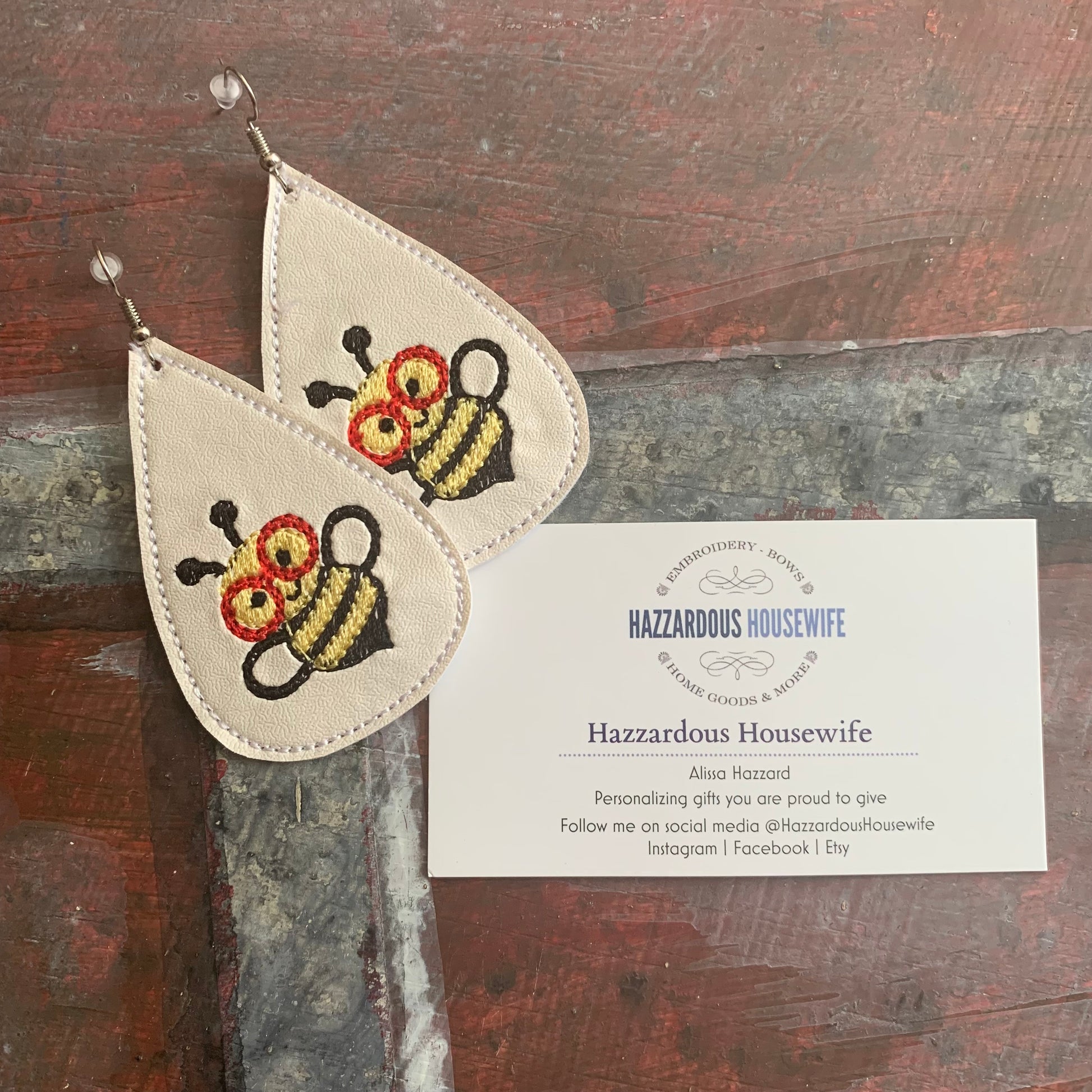 Pair of white earrings with bee designs on a wooden surface, accompanied by a business card for 'Hazzardous Housewife'.