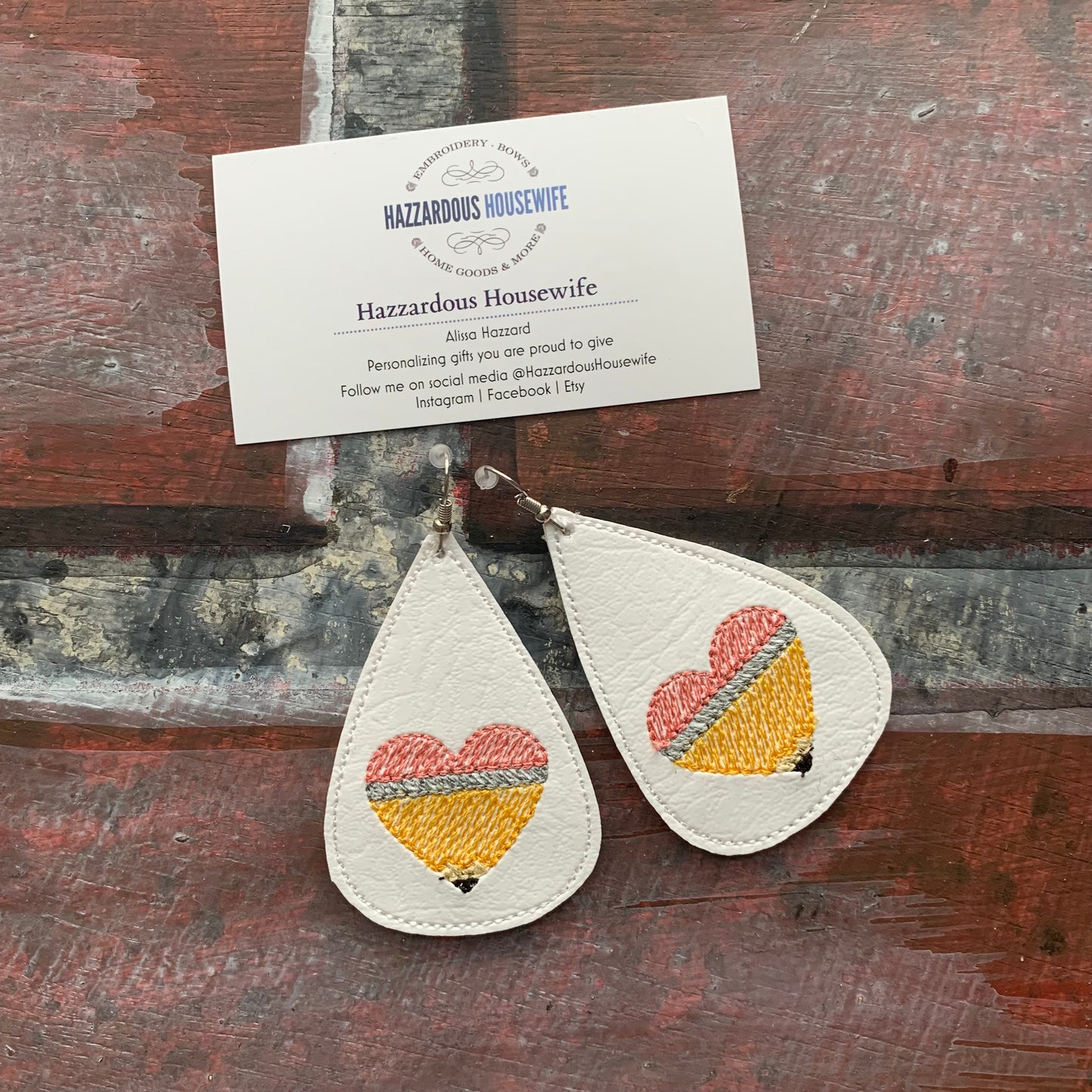 Teardrop-shaped earrings with embroidered hearts on a wooden surface, accompanied by a business card.