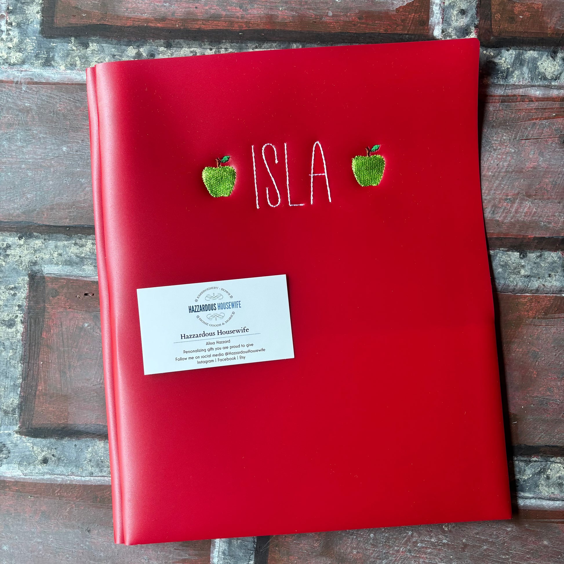 Red plastic folder with embroidered name and 2 green apples on a brick surface