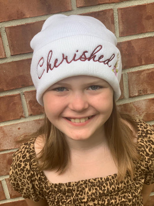 Child wearing a white beanie with 'Cherished' embroidered on it against a brick wall.