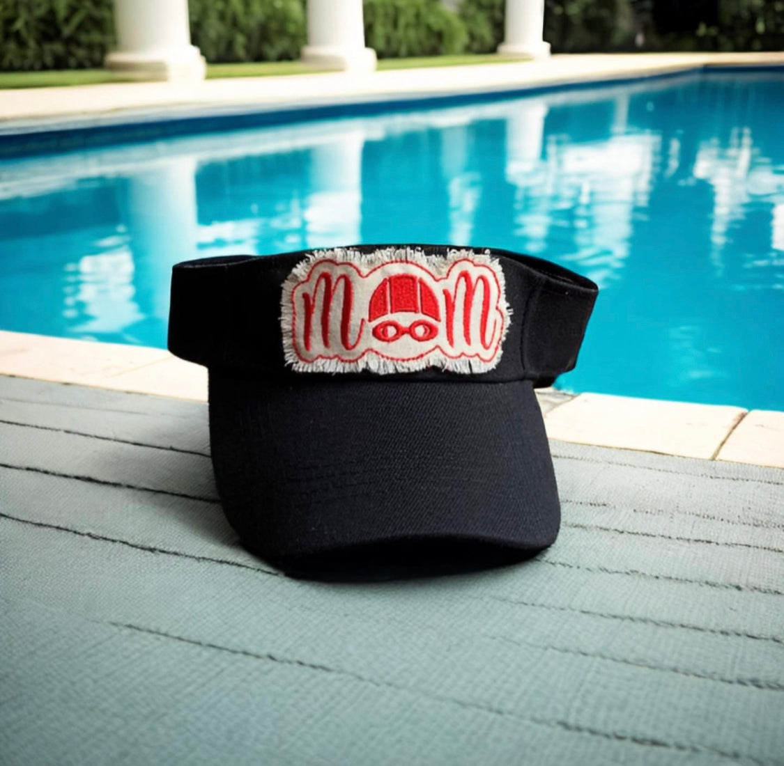 Black visor with a red and white 'mom' patch on a poolside surface