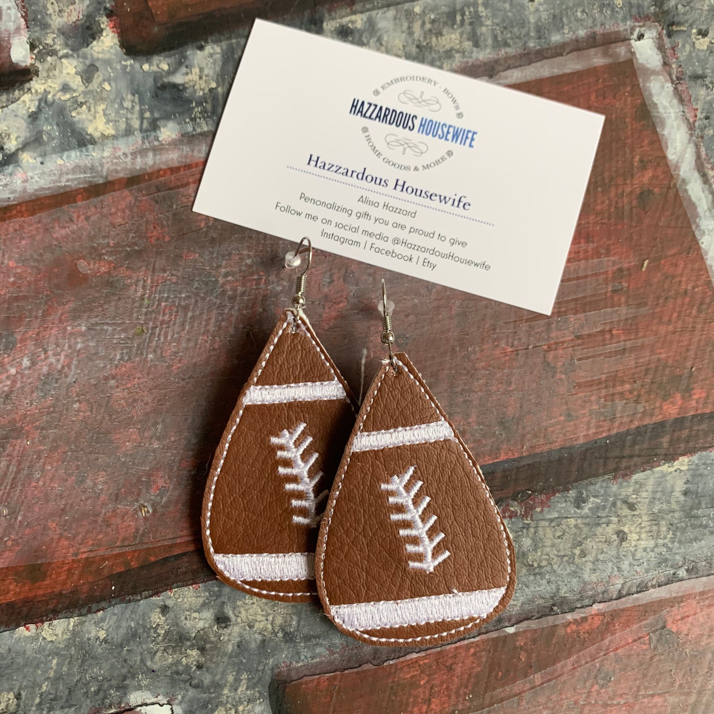 Brown leather football-shaped earrings on a rustic wooden surface with a HazzardousHousewife business card.