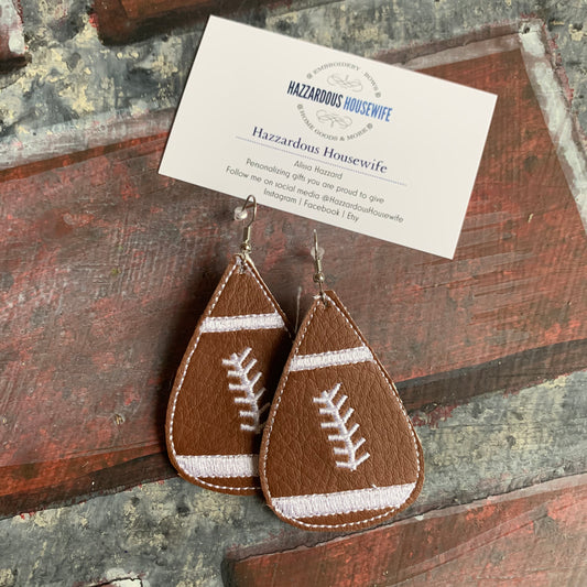 Brown leather football-shaped earrings on a rustic wooden surface with a HazzardousHousewife business card.