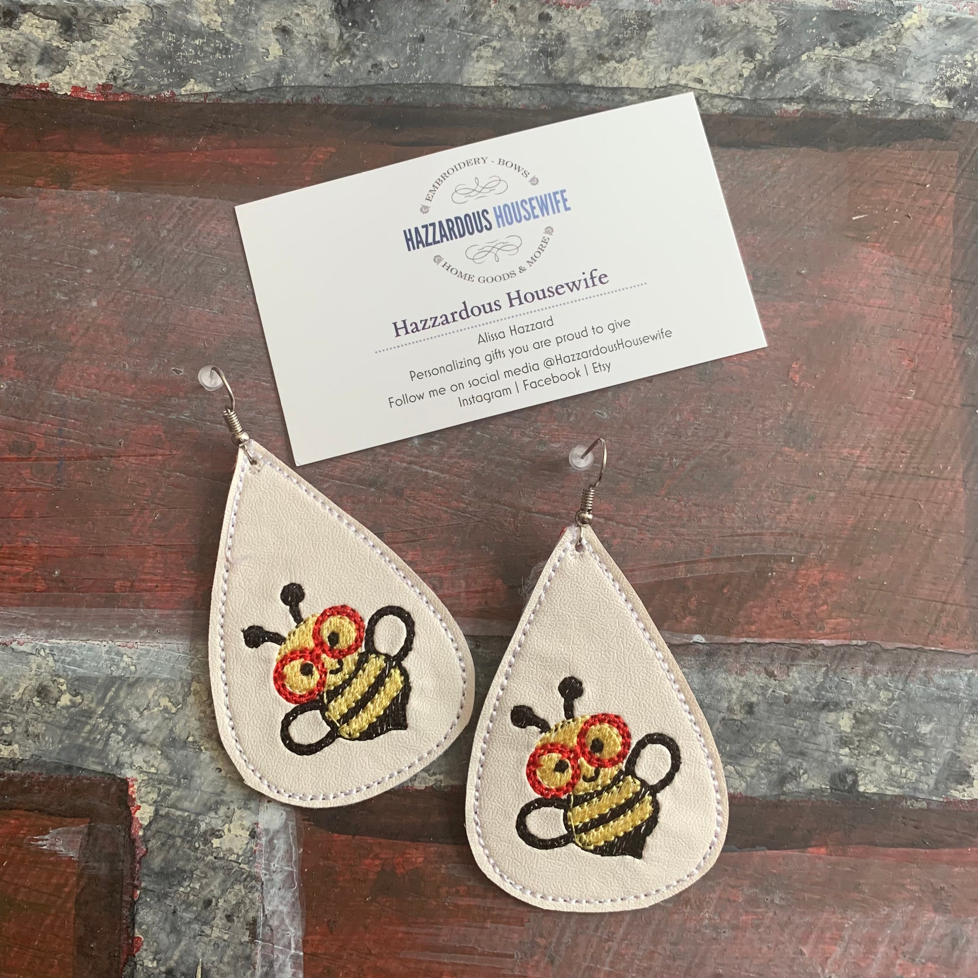 Teardrop-shaped earrings with embroidered bee design on a wooden surface with a business card.