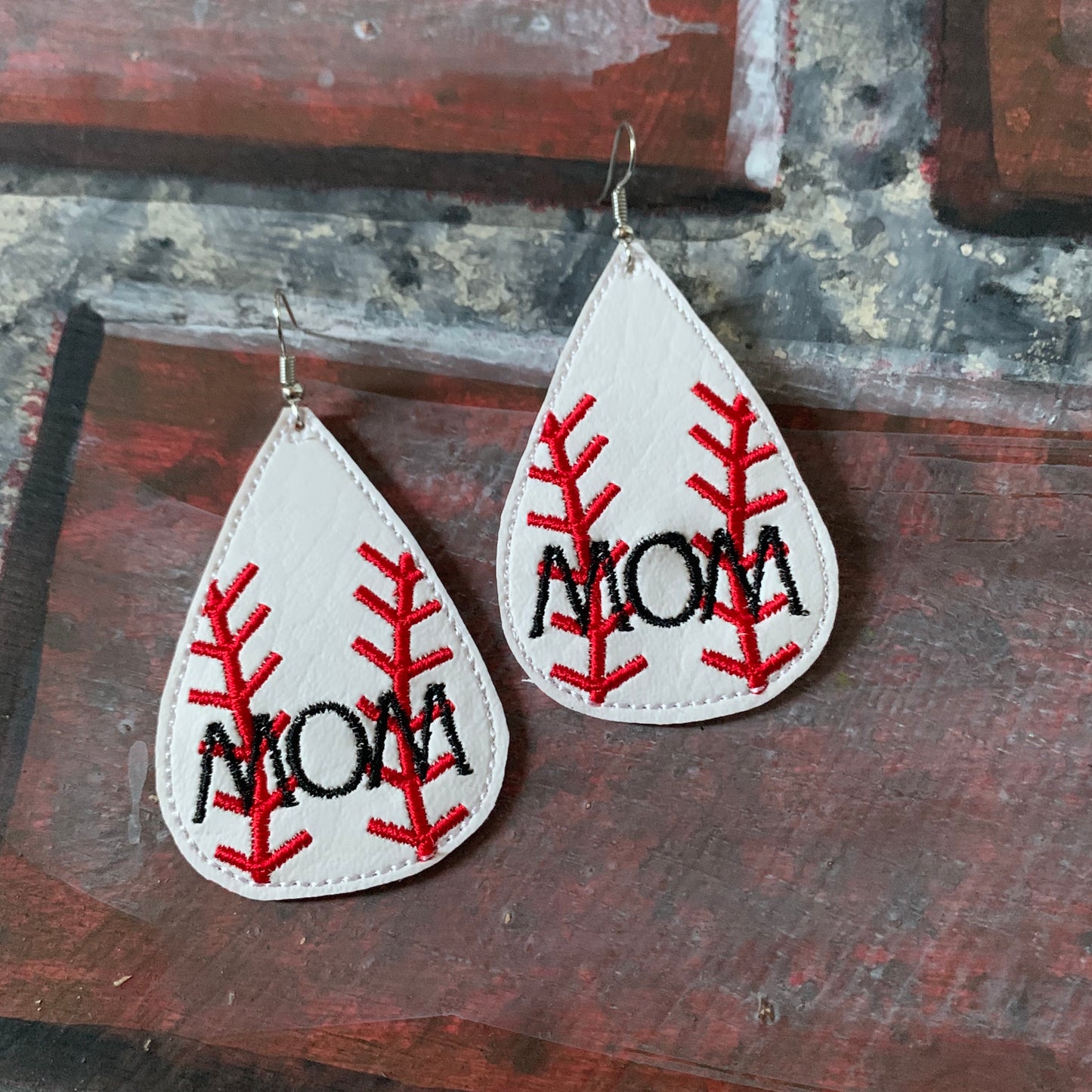 Teardrop-shaped earrings with 'MOM' embroidery on a rustic wooden background