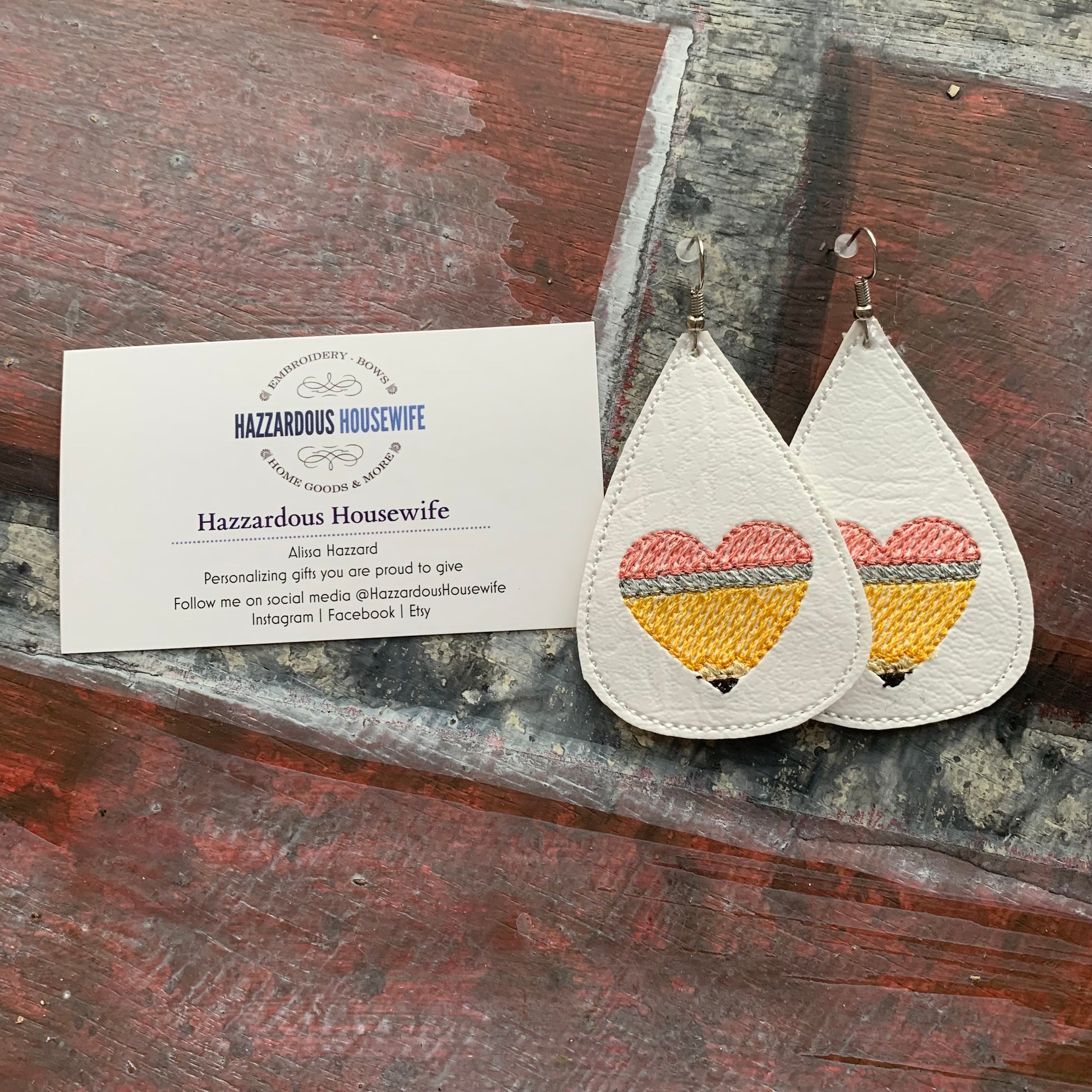 Teardrop-shaped earrings with embroidered pencil in the shape of a heart design on a brick surface with a business card.