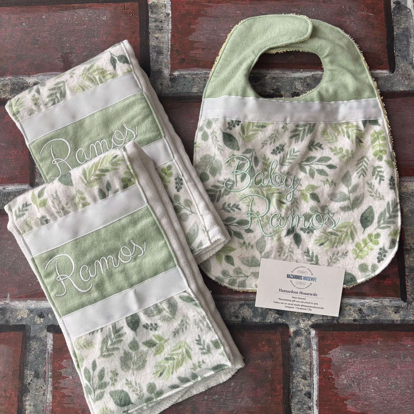 Set of nature green baby bibs and burp cloths with embroidered names on a rustic brick surface.