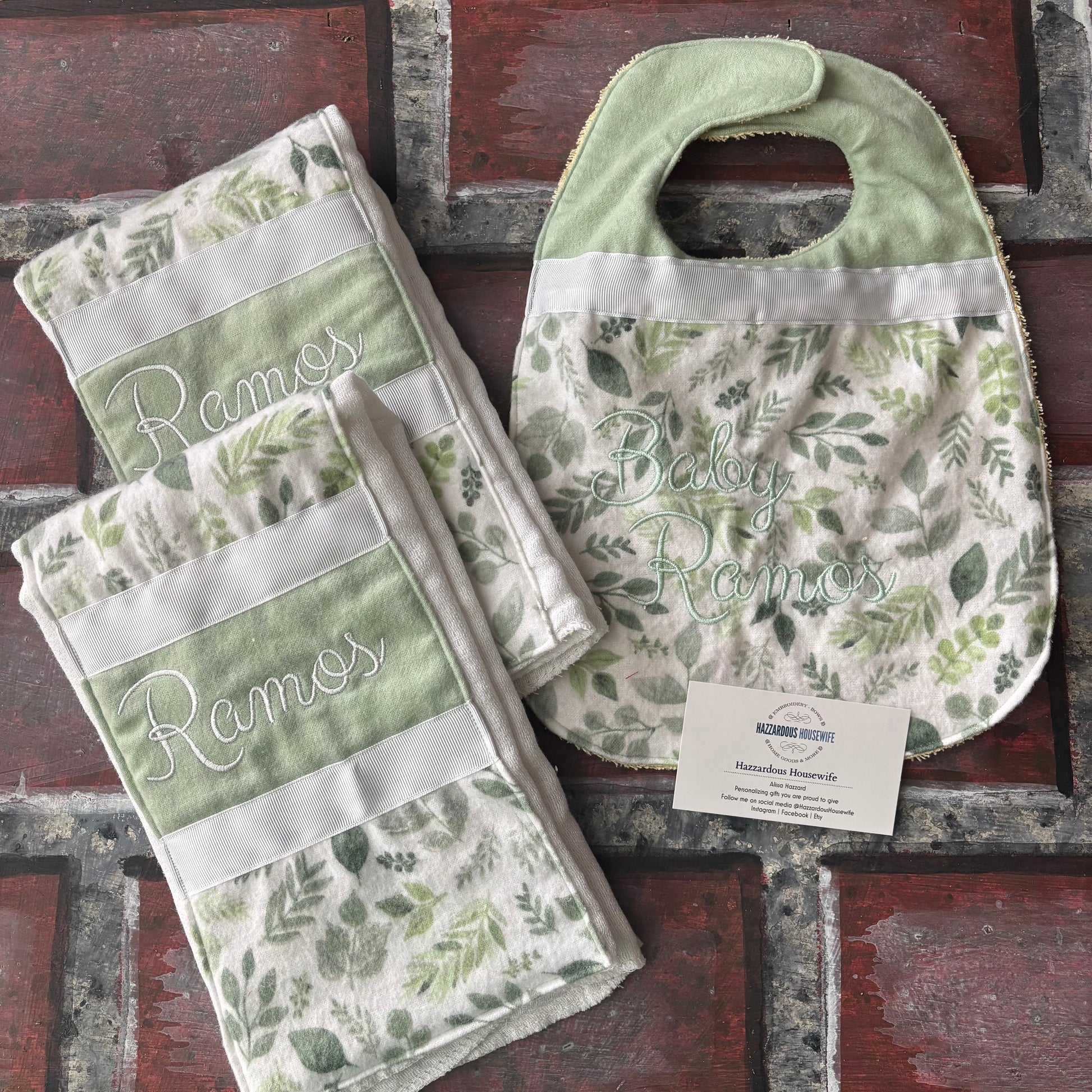 Set of nature green baby bibs and burp cloths with embroidered names on a rustic brick surface.