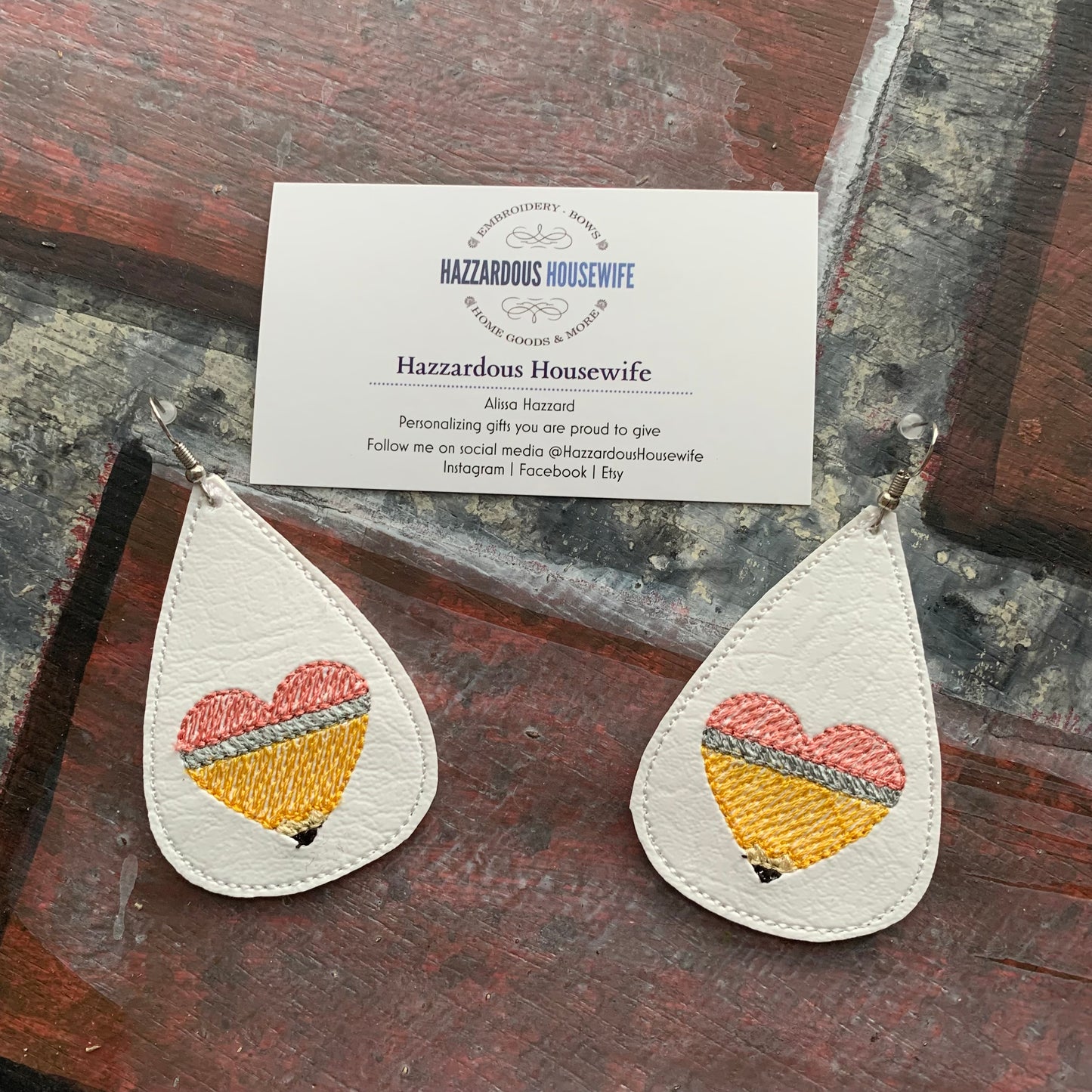 Teardrop-shaped earrings with embroidered pencil in the shape of a heart design on a brick surface with a business card.