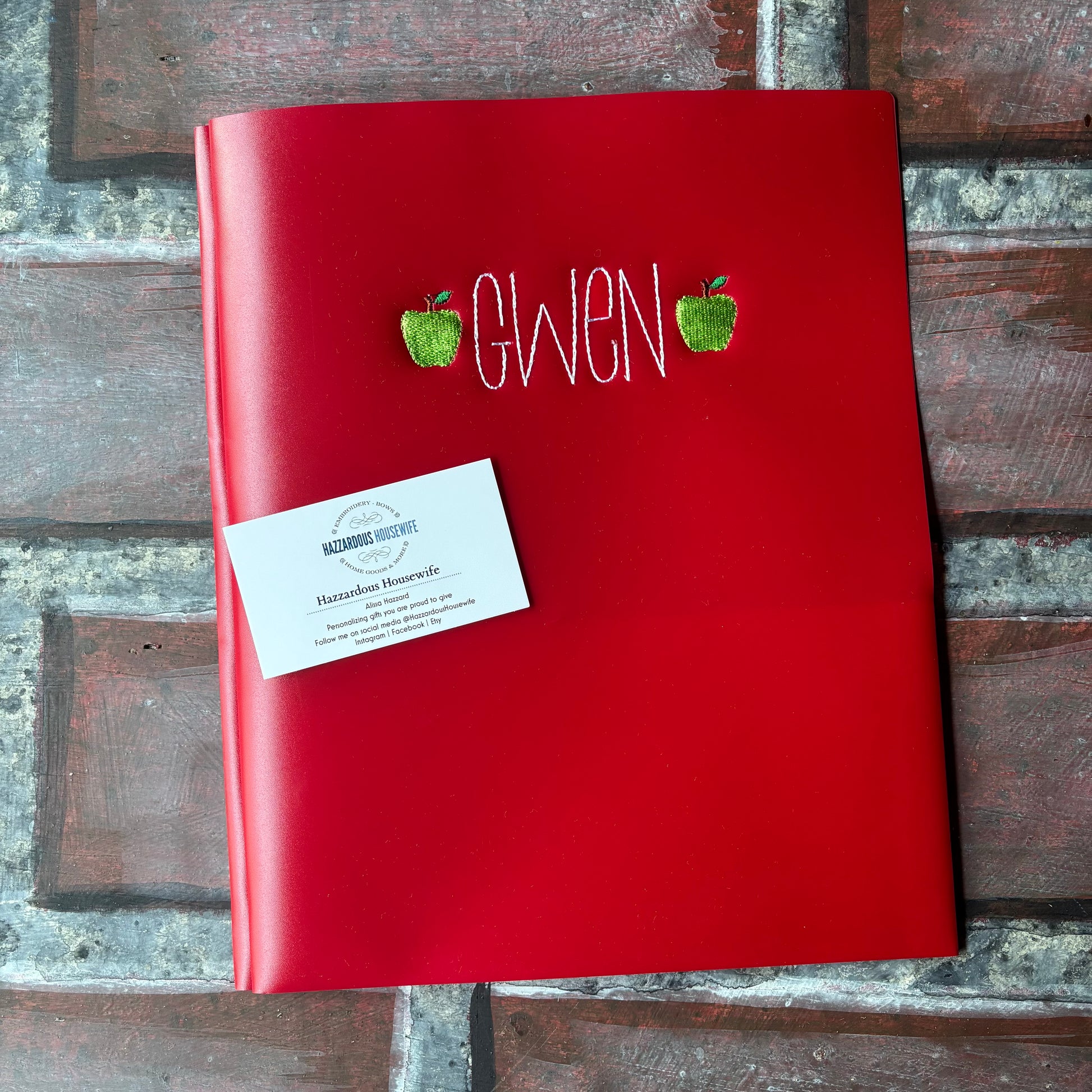 Red notebook folder with 'Gwen' embroidered on it and two green apples, placed on a brick surface.