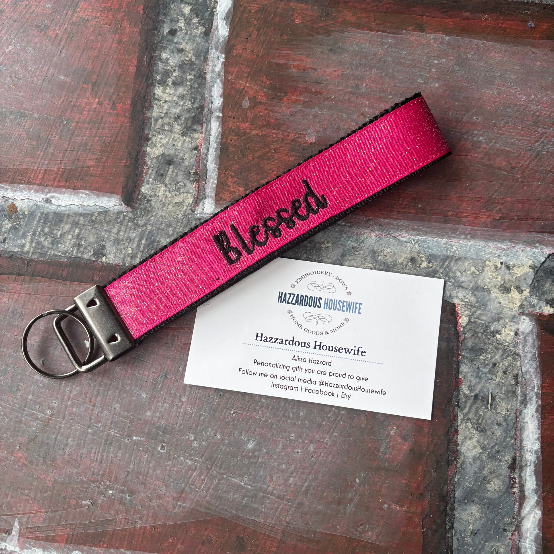 Pink keyfob with black webbing showing at the edge with 'Blessed' embroidered in black text on a wooden surface